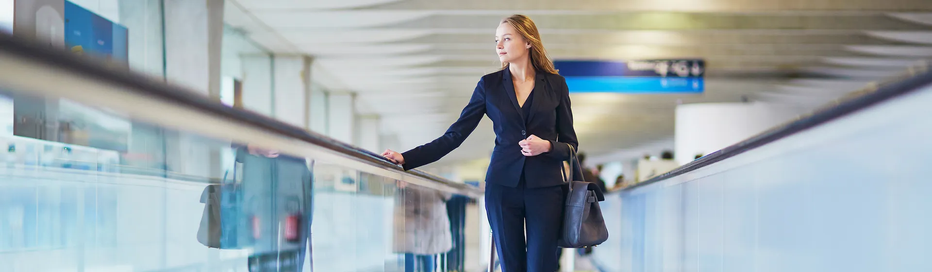 A woman in a business outfit stands on an airport conveyor belt and holds a wheeled suitcase as she gazes relaxed into the distance.