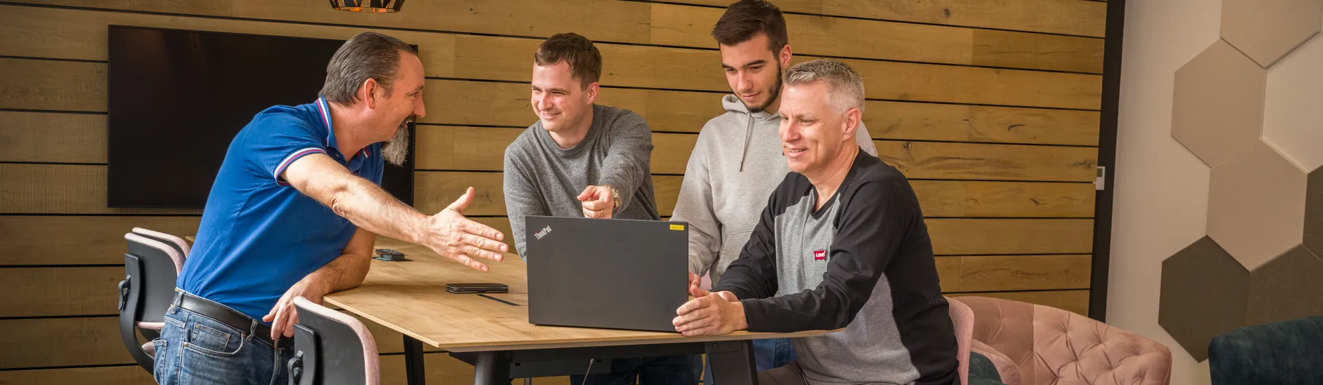 Quatre hommes se tiennent debout autour d’une table haute avec un ordinateur portable dans un bureau moderne et confortable avec des murs en bois et des lampes élégantes.