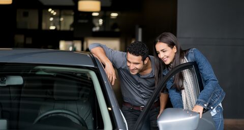 Ein junges Paar steht vor der offenen Tür eine grauen Autos und schauen interessiert hinein.