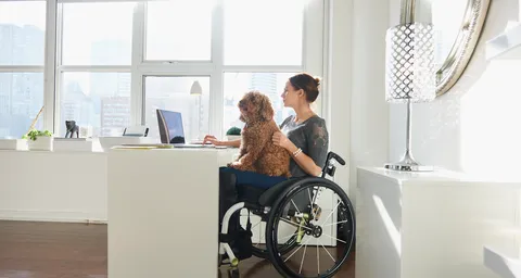 A woman in a wheelchair works on a laptop in a bright, modern room while a small brown dog sits on her lap.
