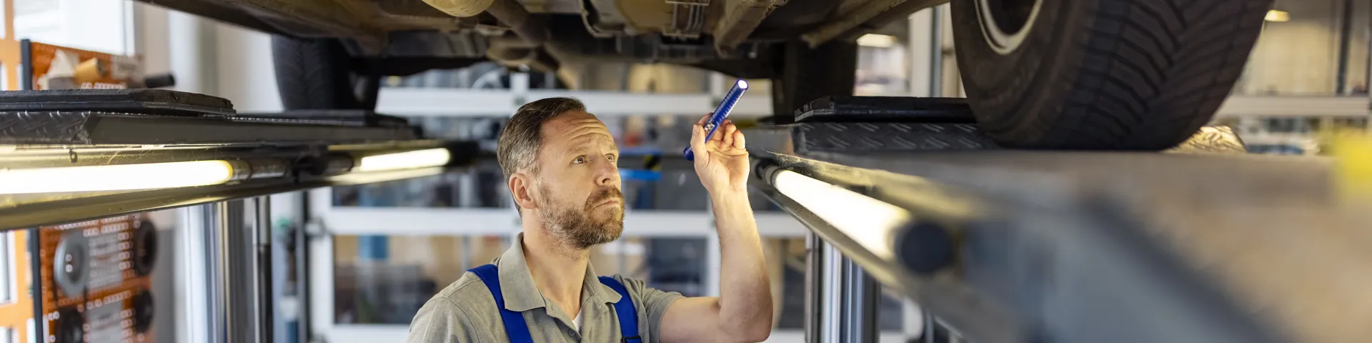 Ein Mechaniker in blauer Latzhose inspiziert ein angehobenes Auto in einer Werkstatt und notiert Details auf einem Tablet.