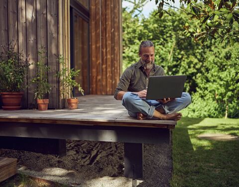 An elderly man sits cross-legged on the porch of his house and looks at his laptop.