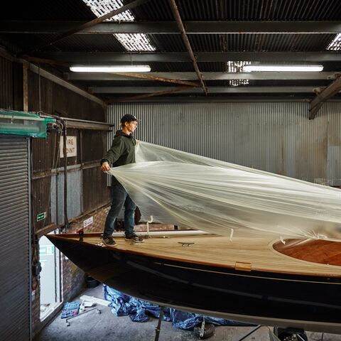 Ein Mann in einer Werkstatt deckt ein grosses Holzboot mit einer durchsichtigen Plastikfolie ab, um es vor Staub und Schmutz zu schützen.