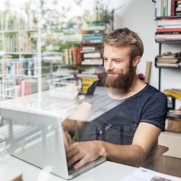 Ein bärtiger Mann arbeitet im Homeoffice am Laptop, mit einer Bücherwand im Hintergrund.