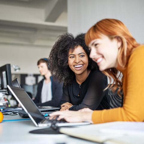 Deux femmes travaillent en riant devant un ordinateur posé sur un bureau.