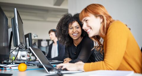 Due donne stanno lavorando al computer in ufficio e ridono.