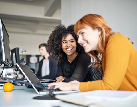 Two women are working on computers in an office and laughing.