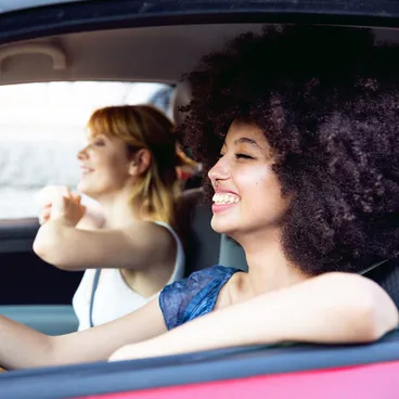 Two cheerful women are sitting in a car, the driver is laughing and the passenger is pointing out of the window, both enjoying the moment.