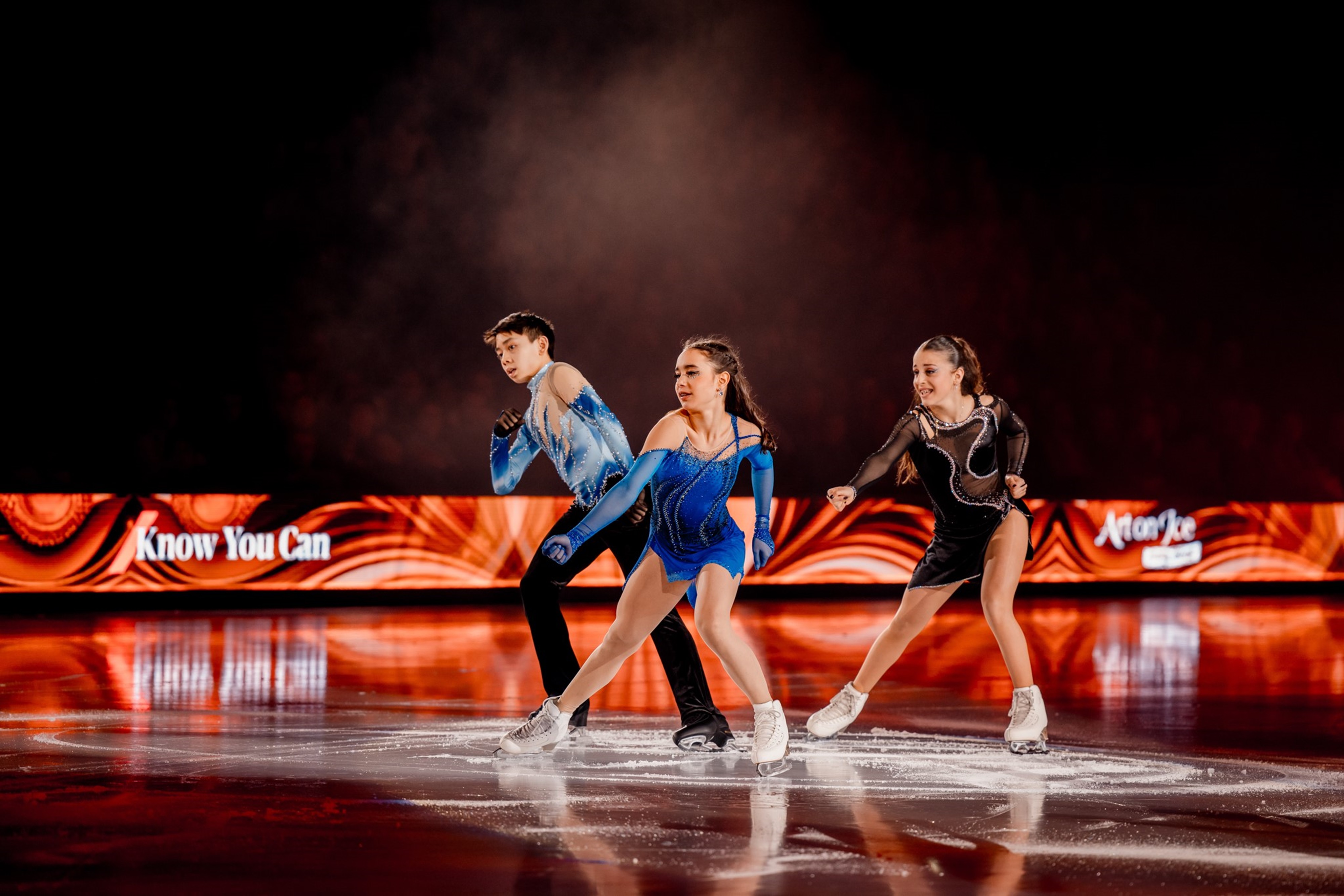 Eine Eiskunstlaufgruppe mit drei Personen in auffälligen Kostümen führt eine Synchron-Performance auf einer beleuchteten Eisfläche vor, mit einer roten Werbebande im Hintergrund.