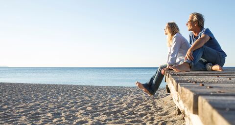 Un uomo e una donna siedono rilassati su una passerella di legno sulla spiaggia e guardano sorridendo il mare sotto un cielo azzurro e limpido.