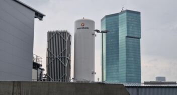 In the foreground is an industrial building with a white tank. In the background is a modern high-rise building with a bluish glow.