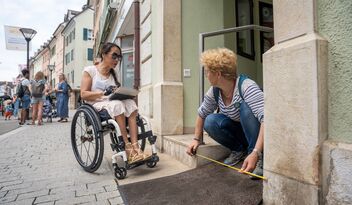 Eine Frau im Rollstuhl und eine weitere Person messen gemeinsam die Höhe einer Eingangsstufe in einer belebten Fussgängerzone.