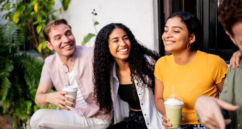 Tre amici sorridenti sono seduti all’aperto su una panchina, immersi nel verde, a bere qualcosa: uno ha un caffè, gli altri un frullato.
