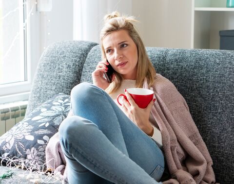 Une femme est assise sur un canapé, elle parle au téléphone et tient une tasse rouge. Elle porte un cardigan rose et a l'air pensif.