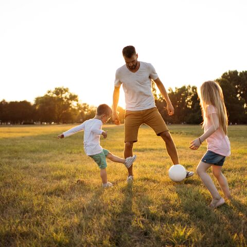 A father is in a large field with his children and is playing football with them.