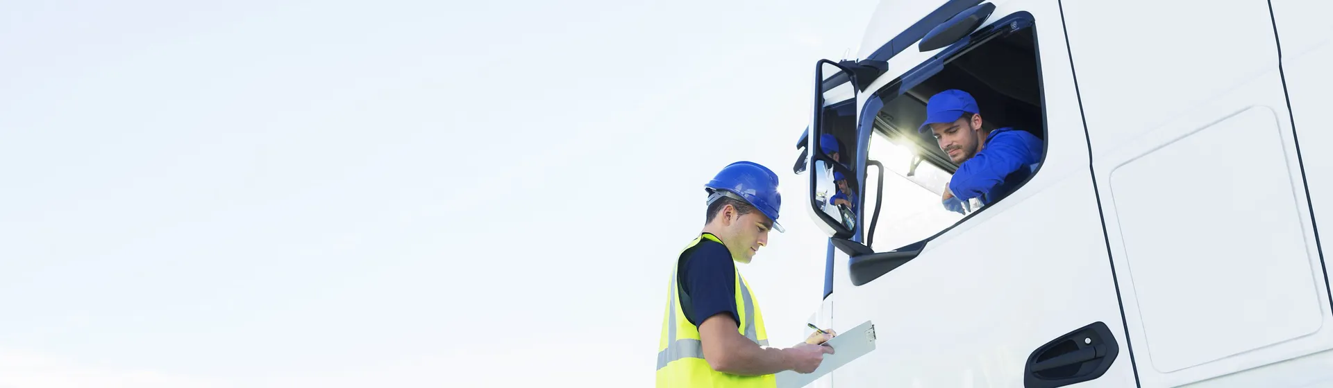 Un ouvrier du bâtiment en gilet de sécurité et casque vérifie des papiers, tandis qu'un chauffeur de camion est assis dans la cabine du camion blanc et observe.