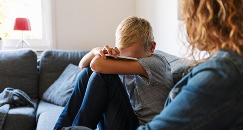 A child sits on a sofa next to a mother, holding his phone and crying.