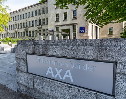 An einer einladenden Mauer prangt der Schriftzug 'Willkommen bei der AXA'. Im Hintergrund erhebt sich ein imposantes Gebäude mit zahlreichen Fenstern, während rechts neben dem Eingang das AXA-Logo sichtbar ist.