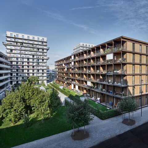 Immeubles d'habitation modernes de plusieurs étages avec des éléments en bois et des balcons, entourés d'espaces verts et d'autres bâtiments en arrière-plan.