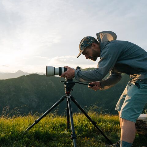 Un homme en équipement de tracking se tient dans une verte prairie et prend une photo avec son appareil.L'appareil photo est posé sur un trépied et on voit les collines en arrière-plan.