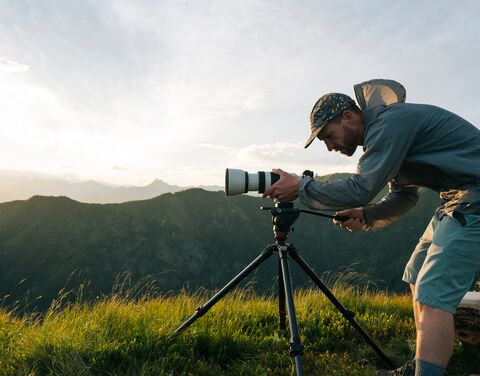 Un uomo è chino sulla sua fotocamera reflex appoggiata su un treppiede sull’erba.