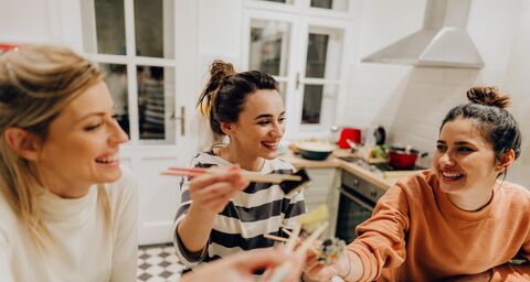 Drei lächelnde junge Frauen sind in der Küche und essen Sushi mit Stäbchen.