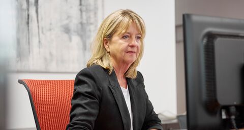 An older woman with medium-length blond hair sits at her desk in the office wearing a black blazer, looking intently at the screen.