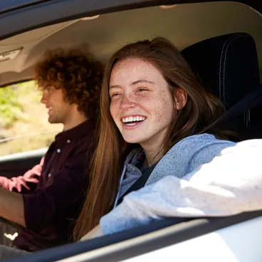 A smiling woman sits in the passenger seat of a car while a man with curly hair drives the car.