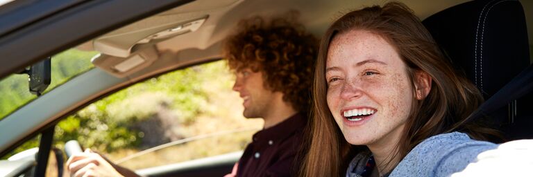 A smiling woman sits in the passenger seat of a car while a man with curly hair drives the car.