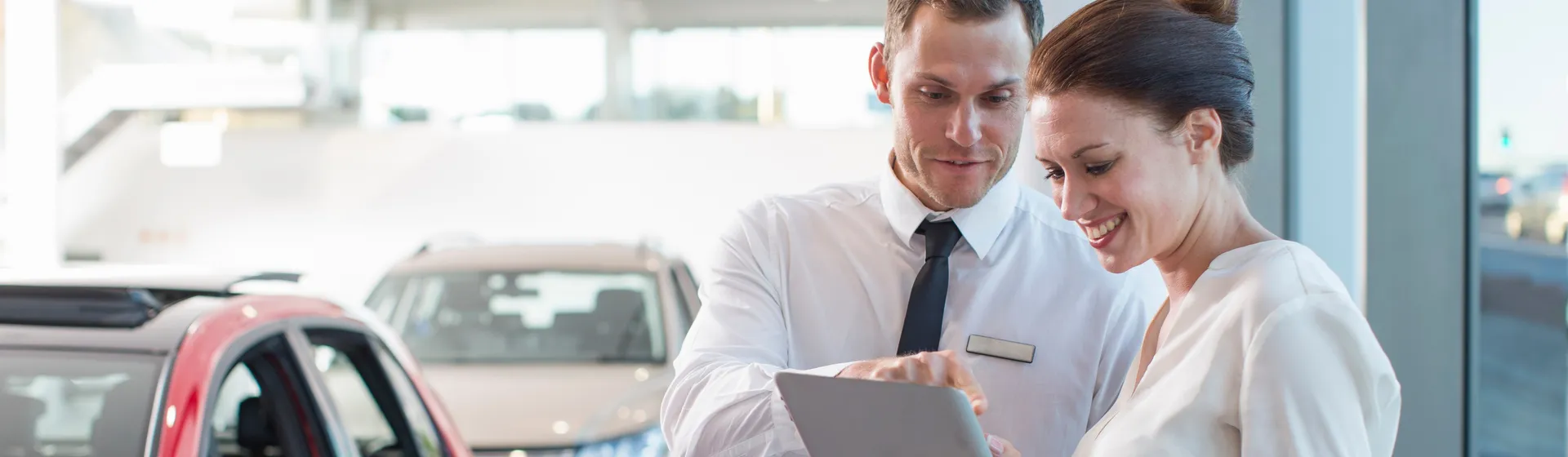 Ein Autoverkäufer im Anzug zeigt einer lächelnden Kundin in weisser Kleidung auf einem Tablet Informationen in einem Autohaus.