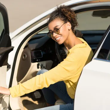 Eine junge Frau mit Sonnenbrille und gelber Jacke steigt lachend aus ihrem weissen Auto.