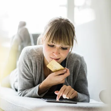 Une femme allongée sur son canapé tient une carte de crédit dans la main et effectue des achats en ligne sur une tablette.