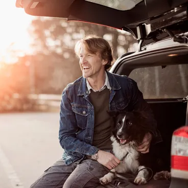 Ein Mann sitzt lächelnd mit seinem Border Collie unter der offenen Heckklappe seines Kombis, während die Sonne im Hintergrund untergeht.